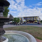 L'image montre une fontaine au premier plan, avec de l'eau qui s'écoule. En arrière-plan, on aperçoit un bâtiment coloré portant le nom "MUSW", entouré de verdure. Le ciel est légèrement nuageux et des personnes se déplacent autour de la fontaine et du bâtiment. L'ensemble donne une impression de vie et d'animation dans un espace public.
