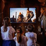 L'image montre un groupe de personnes, probablement une famille, regardant un écran de télévision. Sur l'écran, on peut voir deux personnages, probablement issus d'un jeu vidéo ou d'un film, en pleine discussion. Les quatre personnes qui regardent sont concentrées sur l'écran, avec des expressions intéressées. La pièce est décorée de manière rustique, avec un mur en fourrure derrière la télévision. Les enfants portent des chemises blanches, et une adulte, peut-être leur mère, est à côté d'eux.