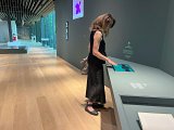 L'image montre une femme debout dans un espace d'exposition, probablement dans un musée. Elle est vêtue d'un haut noir et d'un pantalon long noir. Elle interagit avec une tablette ou un écran tactile placé sur une table, regardant attentivement ce qui s'affiche. L'intérieur de la pièce est moderne, avec des murs clairs, un sol en bois, et plusieurs œuvres d'art visibles en arrière-plan. L'éclairage est naturel, apporté par des fenêtres, créant une ambiance accueillante.