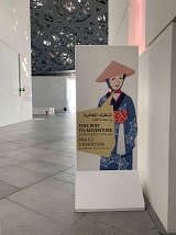 L'image montre un panneau dans un espace intérieur, probablement dans un musée. Le panneau présente une illustration d'une personne vêtue d'un costume traditionnel japonais, avec un chapeau et des motifs décoratifs. À côté de l'image, il y a du texte en plusieurs langues, indiquant la direction vers une aventure au musée des enfants. Le fond est constitué de murs modernes et d'un sol en pierres.