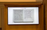 L'image montre un écran où est affiché un livre ouvert. Le texte est écrit dans une calligraphie élégante, probablement en français, mais il est difficile de lire précisément ce qui est écrit. En haut de l'écran, il est mentionné "115 mots sur 20407", indiquant probablement un compte de mots dans un texte plus long. L'arrière-plan semble être en bois, ce qui ajoute une touche chaleureuse à la présentation.