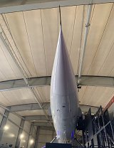 L'image montre un vaisseau ou un modèle de fusée vu de l'avant, se tenant à l'intérieur d'un grand hangar. On peut voir la pointe aérodynamique et lisse du véhicule, qui est en position verticale. L'environnement semble futuriste, avec des éclairages et des structures métalliques visibles autour. La lumière dans l'espace est douce, créant une atmosphère particulière.