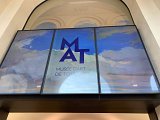 L'image montre une grande affiche située à l'entrée du Musée d'Art de Toulon. L'affiche présente le logo "MAT" en lettres majuscules, sur un fond représentant un ciel avec des nuages. Les couleurs sont douces, dominées par des tons de bleu, évoquant une ambiance artistique et accueillante. L'affiche indique clairement le nom du musée, soulignant son importance culturelle.