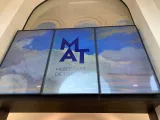 L'image montre une grande affiche située à l'entrée du Musée d'Art de Toulon. L'affiche présente le logo "MAT" en lettres majuscules, sur un fond représentant un ciel avec des nuages. Les couleurs sont douces, dominées par des tons de bleu, évoquant une ambiance artistique et accueillante. L'affiche indique clairement le nom du musée, soulignant son importance culturelle.