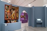 L'image montre une exposition dans un musée, avec un ensemble d'œuvres d'art. À gauche, un retable est présenté, affichant des sculptures et des figures peintes, typiques de l'art religieux. Ce retable est ouvert, révélant plusieurs panneaux avec des scènes, probablement bibliques, et des dorures. En arrière-plan, on peut voir des murs de couleurs différentes, dont un violet, qui contiennent d'autres œuvres ou panneaux d'exposition. Une tablette numérique est également visible sur un support, indiquant des informations supplémentaires. L'ensemble de l'espace est bien éclairé et moderne, créant une atmosphère contemplative pour les visiteurs.