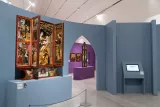 L'image montre une exposition dans un musée, avec un ensemble d'œuvres d'art. À gauche, un retable est présenté, affichant des sculptures et des figures peintes, typiques de l'art religieux. Ce retable est ouvert, révélant plusieurs panneaux avec des scènes, probablement bibliques, et des dorures. En arrière-plan, on peut voir des murs de couleurs différentes, dont un violet, qui contiennent d'autres œuvres ou panneaux d'exposition. Une tablette numérique est également visible sur un support, indiquant des informations supplémentaires. L'ensemble de l'espace est bien éclairé et moderne, créant une atmosphère contemplative pour les visiteurs.