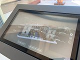 L'image montre un écran interactif, probablement situé dans un musée ou une galerie. Cet écran présente une maquette ou une illustration d'un bâtiment, entourée de divers éléments d'information. On y voit des options de langue, incluant le français (FR), l'anglais (EN) et l'italien (IT). Les termes comme "Histoire animée" et "Histoire des collections" indiquent que l'utilisateur peut explorer des contenus éducatifs ou historiques liés à la collection présentée. L'environnement semble moderne, avec une vue sur un bassin d'eau à l'extérieur.