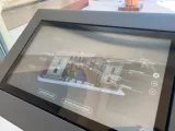 L'image montre un écran interactif, probablement situé dans un musée ou une galerie. Cet écran présente une maquette ou une illustration d'un bâtiment, entourée de divers éléments d'information. On y voit des options de langue, incluant le français (FR), l'anglais (EN) et l'italien (IT). Les termes comme "Histoire animée" et "Histoire des collections" indiquent que l'utilisateur peut explorer des contenus éducatifs ou historiques liés à la collection présentée. L'environnement semble moderne, avec une vue sur un bassin d'eau à l'extérieur.