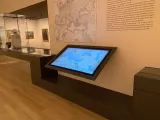 L'image montre un espace d'exposition dans un musée. Au premier plan, il y a un grand écran tactile en forme de tablette, affichant une carte. En arrière-plan, on peut voir un mur avec une carte ancienne de l'Europe et des textes explicatifs. Le sol est en bois, et la lumière de la pièce donne une ambiance chaleureuse. Des œuvres d'art peuvent également être aperçues dans des vitrines, ajoutant à l'atmosphère de l'exposition.