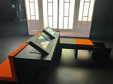 L'image montre un espace intérieur avec des meubles modernes, notamment des tables et des sièges. Les tables sont rectangulaires et possèdent des écrans intégrés, tandis que les bancs sont recouverts d'un tissu orange et noir. L'environnement semble sombre, ce qui donne un aspect contemporain et minimaliste, et il y a un éclairage en arrière-plan qui rappelle des portes ou des fenêtres. Les couleurs dominantes sont le noir et l'orange, créant un contraste visuel marqué.