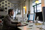 L'image montre un homme assis à un bureau dans un bureau moderne. Il travaille sur un ordinateur avec un écran allumé, et semble concentré sur sa tâche. L'environnement est lumineux, avec des murs clairs et des luminaires au design contemporain. On aperçoit également des plantes en pots et divers objets de bureau disposés sur la table, ce qui contribue à une atmosphère dynamique et professionnelle.