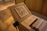 L'image montre un objet en bois, probablement un dispositif interactif ou un meuble design. La surface supérieure présente une gravure d'un animal, peut-être un tigre, entourée d'un cadre en bois. Sur le côté, il y a plusieurs boutons étiquetés "[Fr]", "[En]", et "[De]", suggérant que cet objet pourrait avoir une fonction liée aux langues. En dessous de la surface gravée, des planches en bois sont visibles, indiquant qu'il pourrait s'agir d'un système de rangement ou de présentation. Le sol en bois et l'environnement suggèrent un intérieur moderne et élégant.