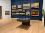 L'image montre une galerie d'art avec des murs peints en bleu. Sur la paroi, plusieurs tableaux sont accrochés, représentant principalement des paysages marins et des scènes de bateaux. Le sol est en bois clair, et au centre de la pièce se trouve une table moderne et basse, avec un écran ou des livres en dessous, invitant les visiteurs à interagir. L'éclairage met en valeur les œuvres d'art au mur.