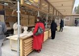 L'image montre un espace de travail en extérieur, structuré avec des échafaudages en bois et en métal. Plusieurs personnes sont occupées à réaliser des activités artisanales, vraisemblablement liées à la sculpture ou à la taille de matériaux. Certaines personnes sont debout derrière des tables où des œuvres ou des éléments en cours de création sont visibles. Une femme en manteau rouge se tient en premier plan, observant ce qui se passe autour d'elle. L'ambiance semble conviviale et créative, et l'environnement est moderne, avec des éléments architecturaux en bois.