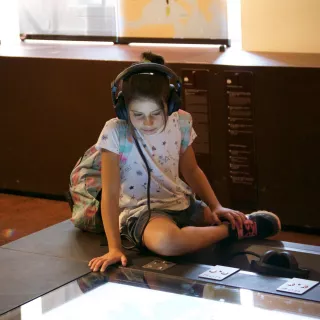 L'image montre une jeune fille assise par terre, absorbée par un écran interactif. Elle porte des écouteurs et regarde attentivement le contenu affiché. Elle est vêtue d'un t-shirt clair avec des motifs et d'un short en jean. À côté d'elle, on peut voir une petite sacoche colorée. L'environnement semble être celui d'un musée, avec des tables d'exposition en arrière-plan.
