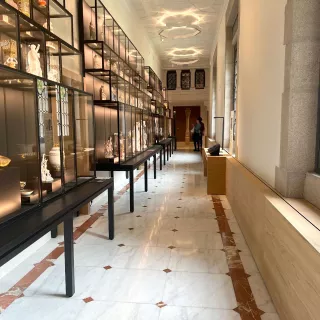 L'image montre un intérieur d'un musée ou d'une galerie. On peut voir une longue allée avec des vitrines en verre noir présentant des objets d'art délicats. Le sol est en marbre clair avec des inserts en marbre rose. À l'arrière-plan, on aperçoit une personne marchant, et de grandes fenêtres laissent entrer la lumière, créant une ambiance lumineuse et accueillante. Les murs sont lisses, et le plafond présente un motif architectural orné.