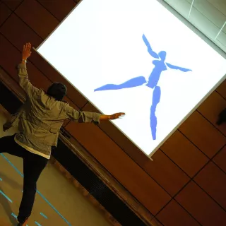 L'image montre une personne en mouvement, peut-être en train de danser ou de faire une performance, se tenant devant un grand écran. Sur cet écran, on voit une silhouette bleue représentant une figure humaine stylisée. L'environnement semble être une salle de réunion ou un espace de présentation, avec des murs en bois et un sol clair. La personne semble interagir avec l'image projetée, ajoutant une dynamique visuelle à la scène.