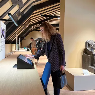 L'image montre une femme se tenant près d'un comptoir dans un musée. Elle semble interagir avec un écran tactile. En arrière-plan, on peut apercevoir d'autres visiteurs et une statue ou une sculpture ancienne sur un socle. La pièce a des poutres en bois au plafond, créant une ambiance chaleureuse et accueillante. La femme porte une tenue professionnelle avec un sac à main.