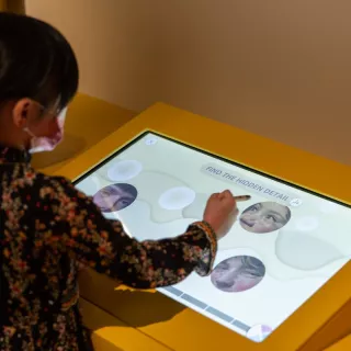 L'image montre une jeune fille utilisant un écran tactile. Elle semble concentrée sur une activité interactive, où elle doit trouver un détail caché. L'écran affiche des visages flous ou modifiés, et la représentation de l'interface utilise des formes fluides. La fille porte un masque et est habillée d'une robe à motifs floraux, ce qui ajoute une touche colorée à la scène. Le décor est minimaliste, avec un fond jaune qui attire l'attention sur l'écran.