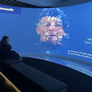 L'image montre une installation interactive dans un espace muséal. À l'avant, une femme est assise sur un banc, regardant une projection immersive sur un mur courbé. La projection affiche une œuvre d'art numérique qui semble déformer un visage, évoquant une expression émotionnelle. À côté de l'image, du texte est visible, mentionnant un soldat dans une zone de déploiement, ce qui suggère un thème lié à la guerre ou à l'histoire militaire, en particulier en rapport avec la Normandie. L'éclairage et le design moderne de l'espace ajoutent une ambiance contemplative à l'ensemble de l'exposition.