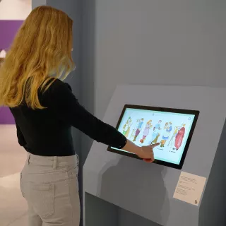L'image montre une personne avec des cheveux longs et blonds, en train d'interagir avec un écran tactile. Elle semble se trouver dans un musée ou une exposition, devant un mur de couleur bleue. Sur l'écran, il y a des illustrations de personnages, probablement liés à un thème culturel ou historique. L'environnement apparaît moderne et épuré.