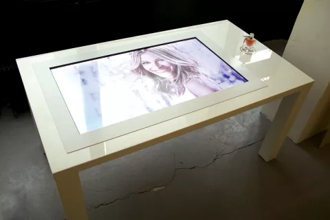 L'image montre une table moderne avec un plateau en verre. Sur ce plateau se trouve un écran tactile affichant une image, probablement d'une personne souriante. À côté de l'écran, il y a un flacon de parfum élégamment placé. La table a un design contemporain, avec des lignes épurées et une finition blanche, et elle est située dans un environnement intérieur minimaliste.