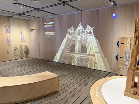 L'image montre un espace intérieur, probablement une exposition. On y voit un mur en bois et une projection d'une façade d'église, mettant en avant des éléments architecturaux comme des arcs et des fenêtres. À côté, il y a des illustrations et des textes explicatifs sur la conception d'une flèche. Le sol semble en bois, et il y a des structures en forme de demi-cercle qui pourraient servir de sièges ou de supports d'exposition. L'ensemble donne une impression moderne et éducative, mêlant art et histoire.