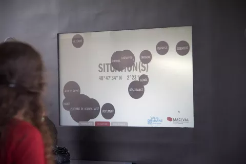 L'image montre un écran affichant le mot "SITUATION(S)" accompagné de plusieurs mots-clés en noir, tels que "IDENTITÉ," "DOCUMENT," et "RÉSISTANCE." Ces mots sont disposés de manière désordonnée, créant une sorte de carte mentale. En arrière-plan, on peut voir une personne dont seule la silhouette est visible, suggérant qu'elle regarde l'écran. L'environnement semble moderne et épuré.