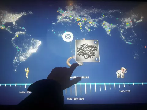 L'image montre un écran interactif représentant une carte du monde, avec des points lumineux qui pourraient indiquer des emplacements géographiques ou des événements. En bas, il y a une frise chronologique qui s'étend de 1500 à 1800, avec des repères temporels. Un doigt est aperçu en train d'interagir avec l'écran, probablement pour sélectionner ou explorer des informations. Des éléments graphiques, comme une sculpture et un éléphant, sont également visibles, indiquant peut-être un lien avec des cultures ou des périodes historiques.