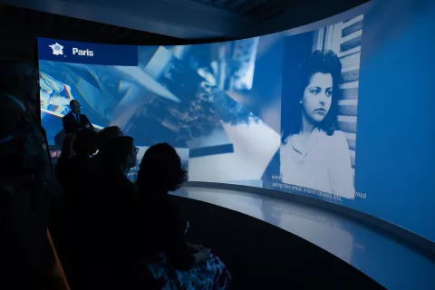 L'image montre une installation artistique ou un événement avec un écran incurvé affichant des contenus visuels. À gauche, une personne semble présenter ou parler, tandis qu'à droite, on voit une photo en noir et blanc d'une femme. Le fond est illuminé et les spectateurs, assis en face, regardent attentivement. Le mot « Paris » est affiché en haut. L'atmosphère semble être celle d'une exposition ou d'une présentation multimédia.