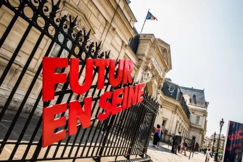 L'image montre un bâtiment, probablement une institution importante, avec un grille en fer forgé. Sur la grille, il y a une inscription en lettres rouges qui dit "FUTUR EN SEINE". En arrière-plan, on aperçoit des gens qui déambulent, et un drapeau est visible sur le toit du bâtiment. Le ciel est clair et ensoleillé, ce qui donne une ambiance sympathique à la scène.