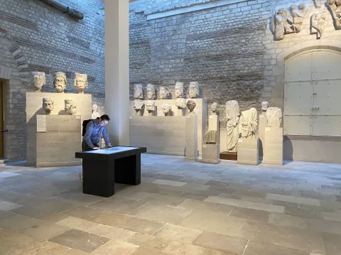 L'image montre une salle d'exposition avec des sculptures anciennes. À gauche, plusieurs bustes en pierre sont disposés sur des socles, tandis qu'à droite, une autre sculpture intrigante est visible. Un homme est penché sur une table noire au centre de la pièce, probablement en train d'examiner un document ou une carte. La lumière naturelle entre à travers des fenêtres, créant une atmosphère calme et contemplative, accentuée par les murs en pierre apparente.