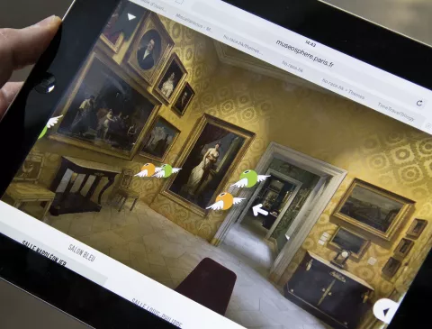 L'image montre une personne tenant une tablette affichant une visite virtuelle d'un musée. Sur l'écran, on peut voir une salle de musée avec des peintures accrochées aux murs, ainsi qu'un mobilier ancien. Les murs sont décorés de motifs et il y a des flèches indiquant les directions à explorer, suggérant une interface interactive pour naviguer dans l'espace.