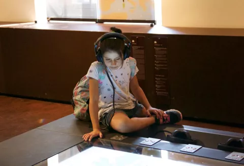 L'image montre une jeune fille assise sur le sol, avec des écouteurs sur les oreilles. Elle porte un t-shirt blanc avec des motifs et un sac à dos coloré. Elle semble concentrée sur un écran interactif devant elle, qui pourrait afficher une carte ou une information. L'environnement est celui d'un musée, avec un éclairage naturel et des panneaux explicatifs en arrière-plan.
