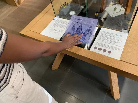 L'image montre une personne en train d'interagir avec un écran tactile placé sur une table en bois. On peut voir une représentation graphique d'un bâtiment, probablement une illustration d'un monument ou d'un lieu historique. À côté de l'écran, il y a des panneaux explicatifs contenant des informations. L'espace semble être une exposition, peut-être dans un musée ou une galerie. L'ensemble de la scène dégage une ambiance éducative et interactive.