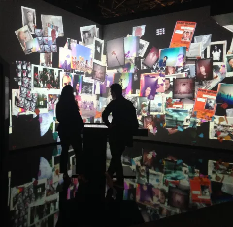 The image depicts a visually immersive installation featuring a large collection of photographs and images projected onto walls. Two silhouettes are visible in the foreground, standing and observing the display. The surrounding images include a variety of photographs, possibly including portraits and artistic visuals, creating a vibrant and dynamic environment. The floor appears reflective, adding to the overall visual complexity of the scene.