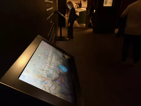 L'image montre un dispositif interactif, probablement un écran tactile, monté sur un support. L'écran affiche une interface numérique, peut-être dédiée à une exposition ou à une présentation. On peut voir du texte et potentiellement des images ou des graphiques sur l'écran. Le fond est sombre, ce qui met en valeur l'écran. À proximité, il y a un panneau explicatif sur le mur, qui pourrait fournir des informations supplémentaires sur le sujet présenté.