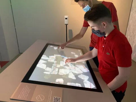 L'image montre deux jeunes garçons en train d'interagir avec un panneau tactile. Ils portent des masques et sont habillés en t-shirts rouges. Le panneau affiche des images de morceaux de pierres ou de fragments, probablement dans un contexte éducatif ou muséal, où ils explorent et découvrent des informations. L'ambiance semble moderne et engageante, favorisant l'apprentissage interactif.